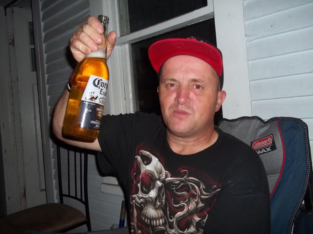 A man sits outside, holding a drink and wearing a relaxed outfit, enjoying the evening atmosphere.