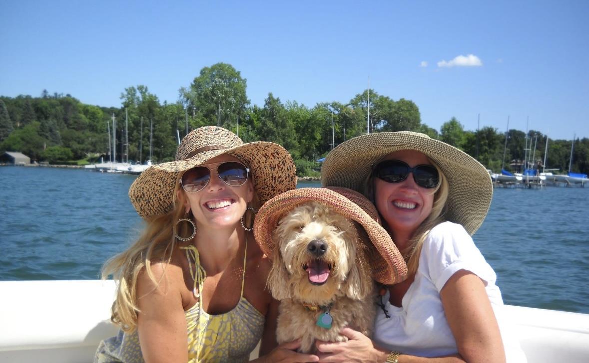 Two women and a dog share joy on a boat, wearing sun hats, under a clear blue sky.