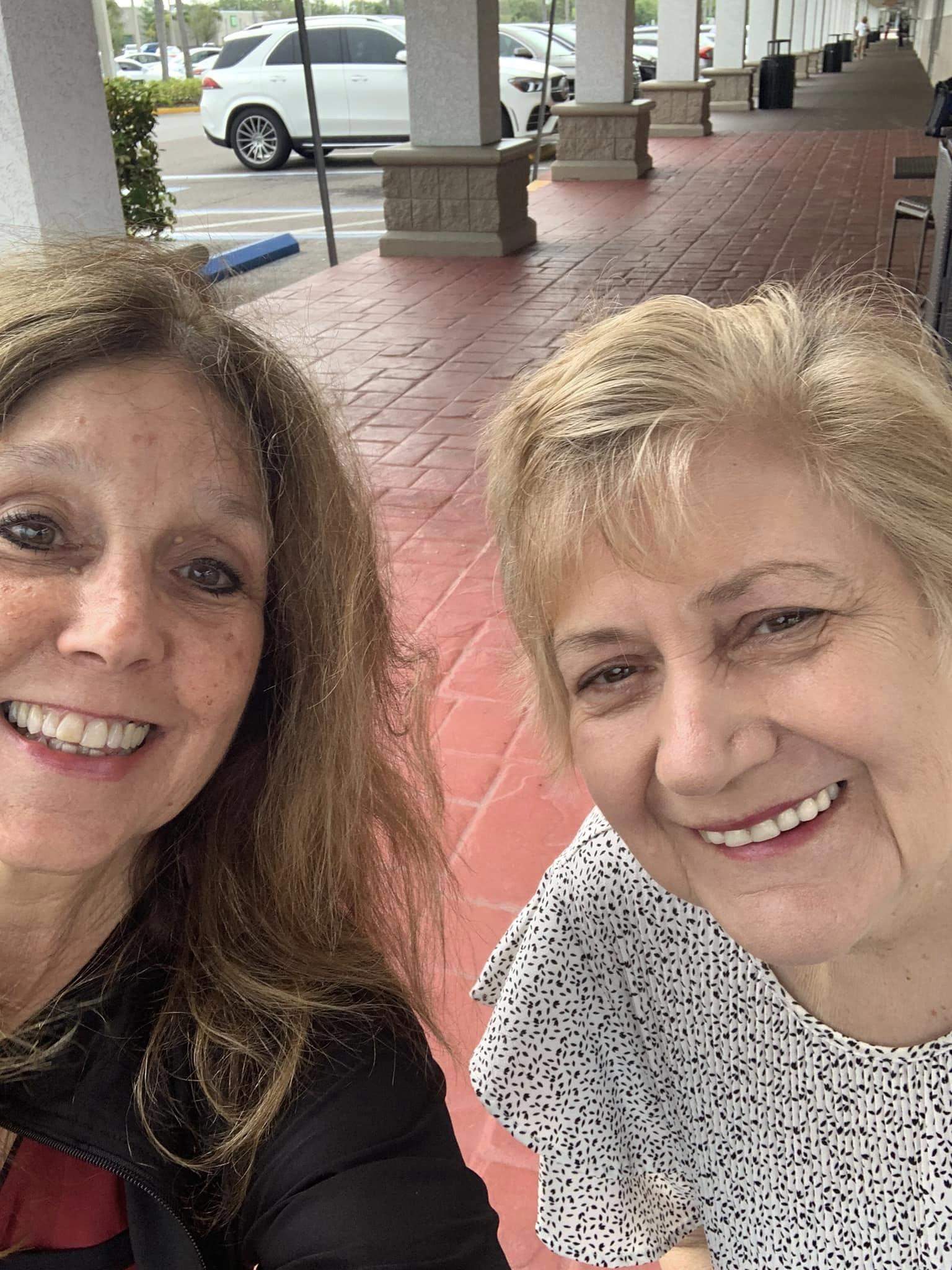 Friends share smiles while taking a selfie outside the storefront on a bright day.