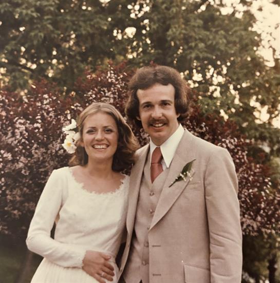 A happy couple stands together in a garden, surrounded by vibrant flowers on a sunny day.