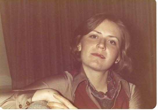 A young woman with curly hair sits comfortably, wearing a fashionable outfit from the 1970s.