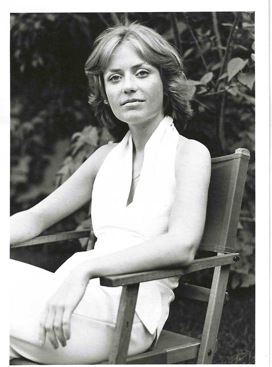A woman with shoulder-length hair sits confidently in a chair surrounded by greenery.