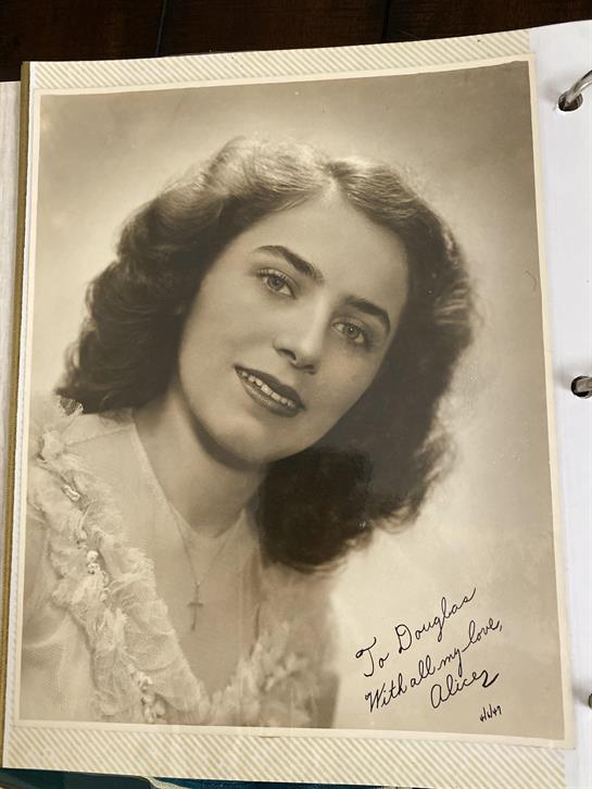 A vintage portrait of a smiling woman in a light dress with curly hair and a cross necklace.