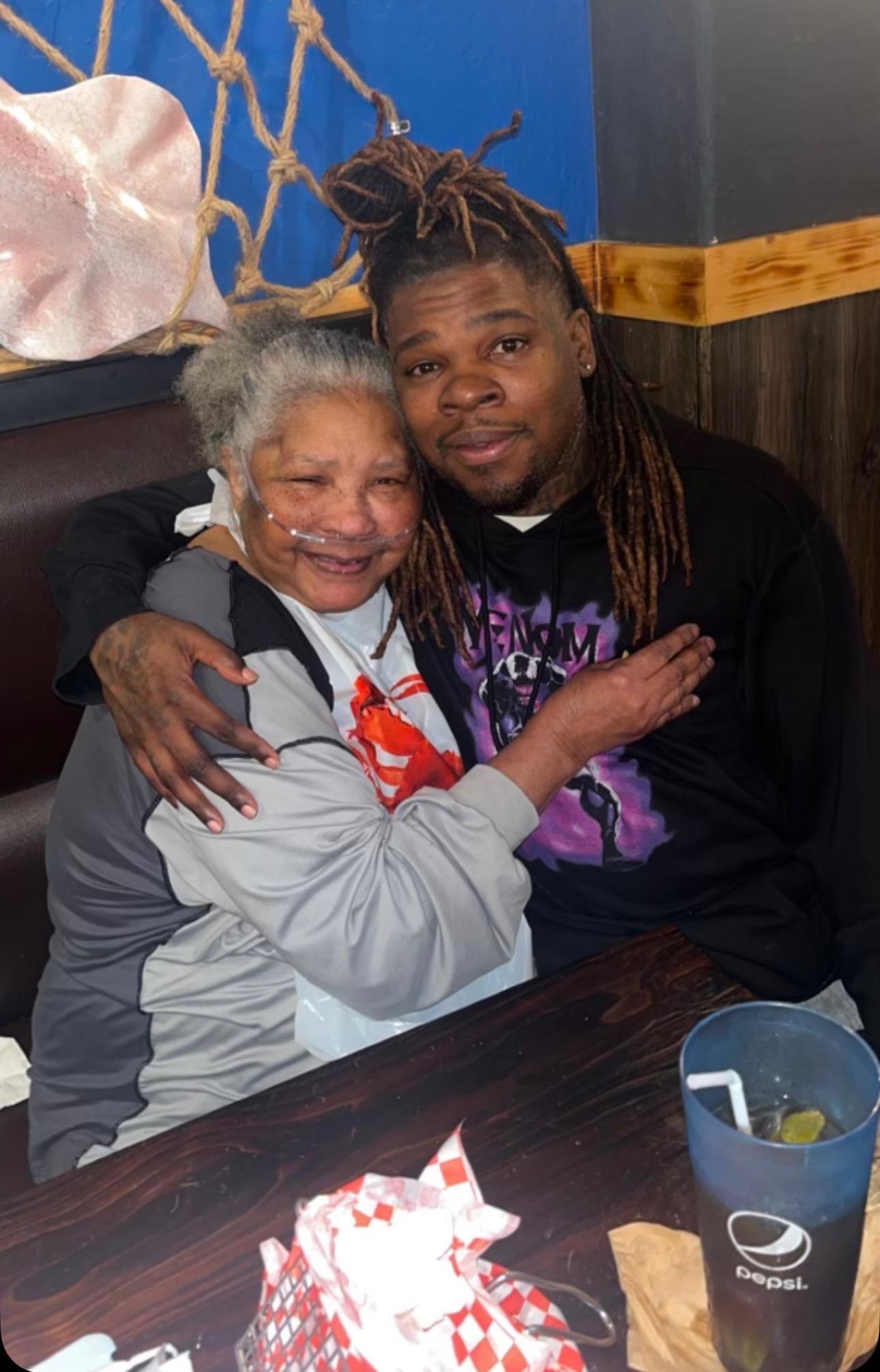 Elderly woman smiles warmly as she embraces a young man in a relaxed dining atmosphere.