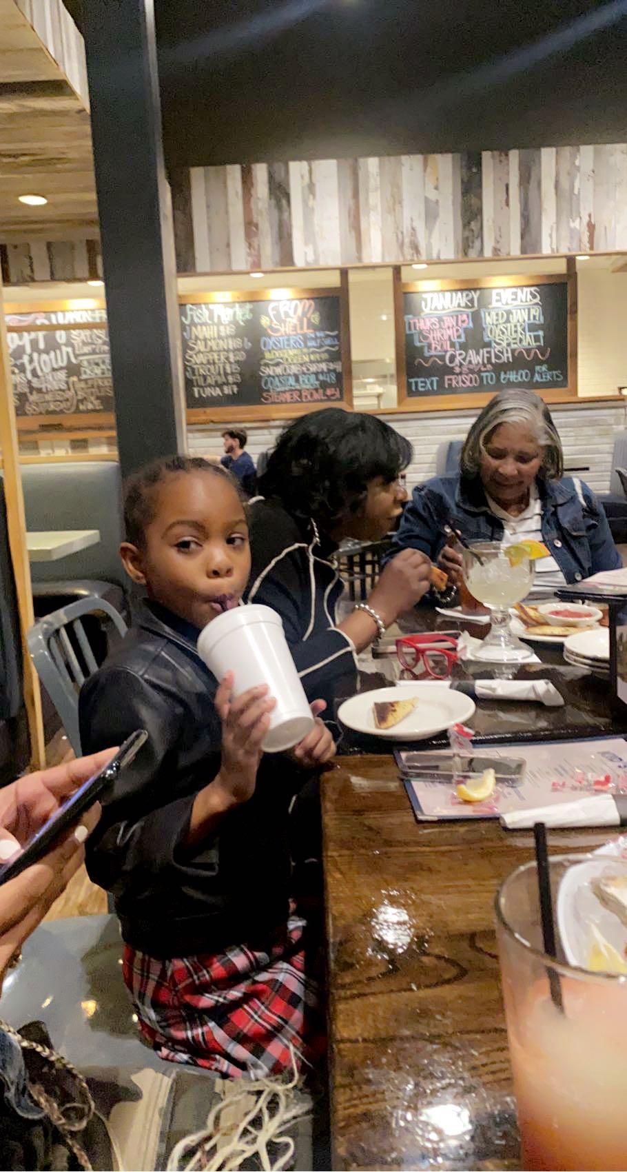A young girl drinks from a cup while family members eat and chat at a restaurant table.