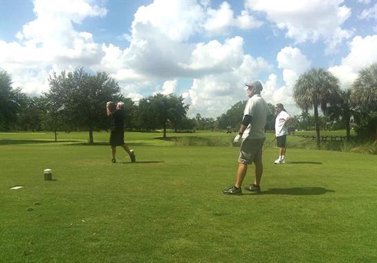 Golfers take turns teeing off on a scenic golf course under a bright blue sky with clouds.