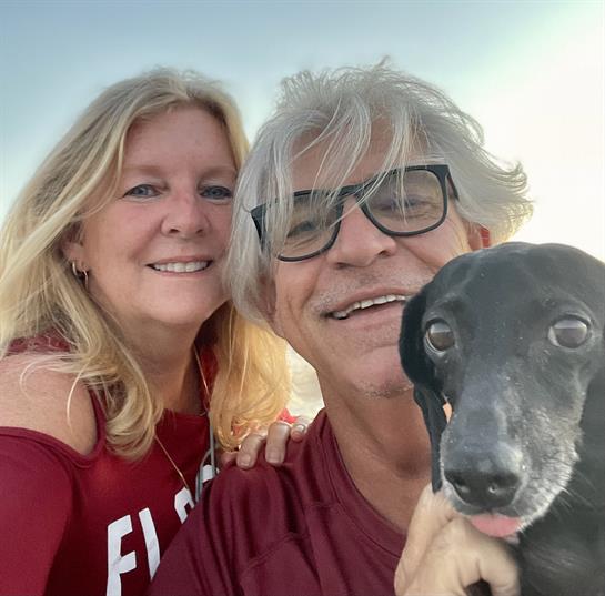 A couple smiles widely while posing with their dog on the beach during sunset.