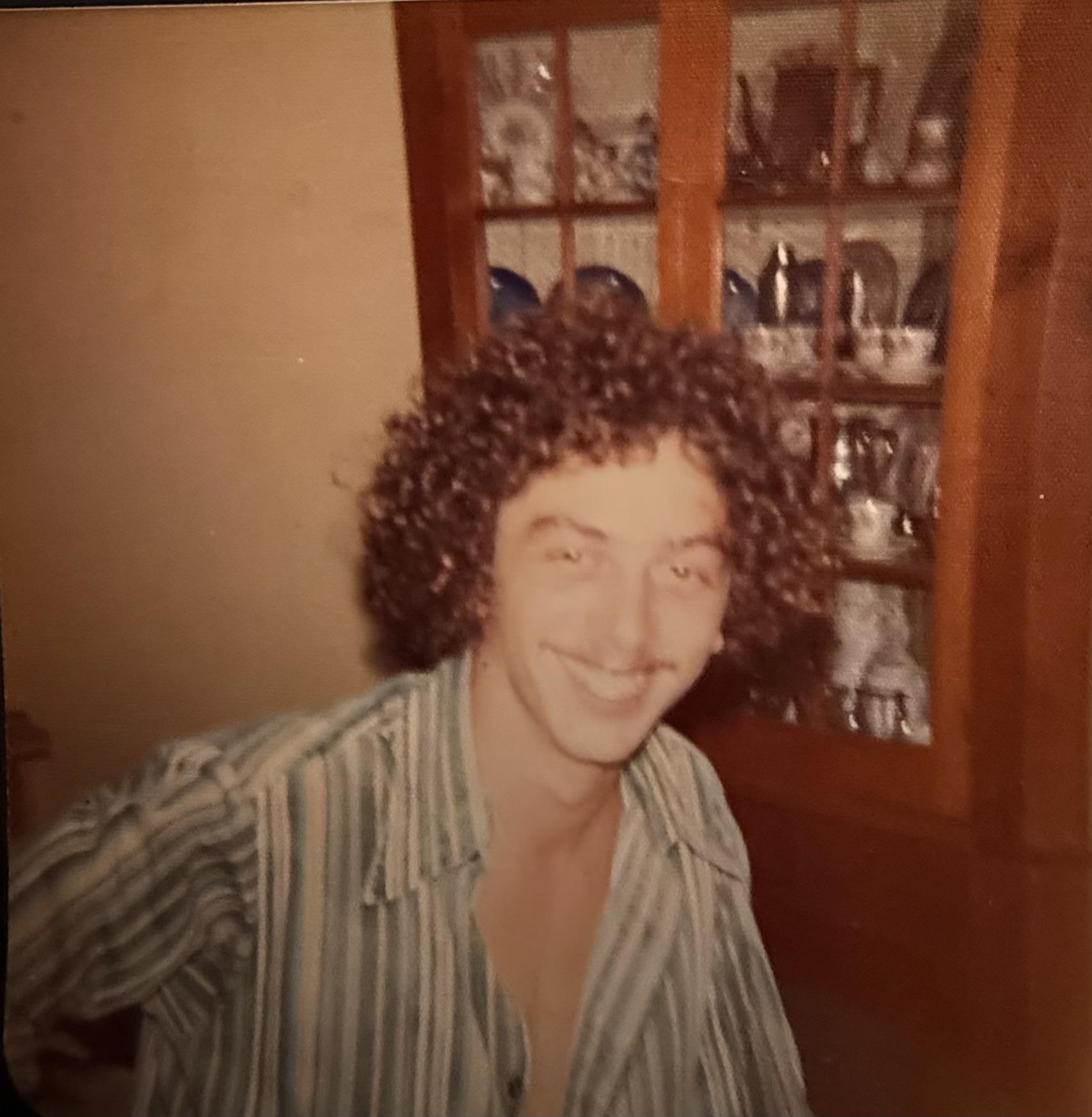 A young man smiles broadly while sitting in a cozy vintage room with a wooden display cabinet.