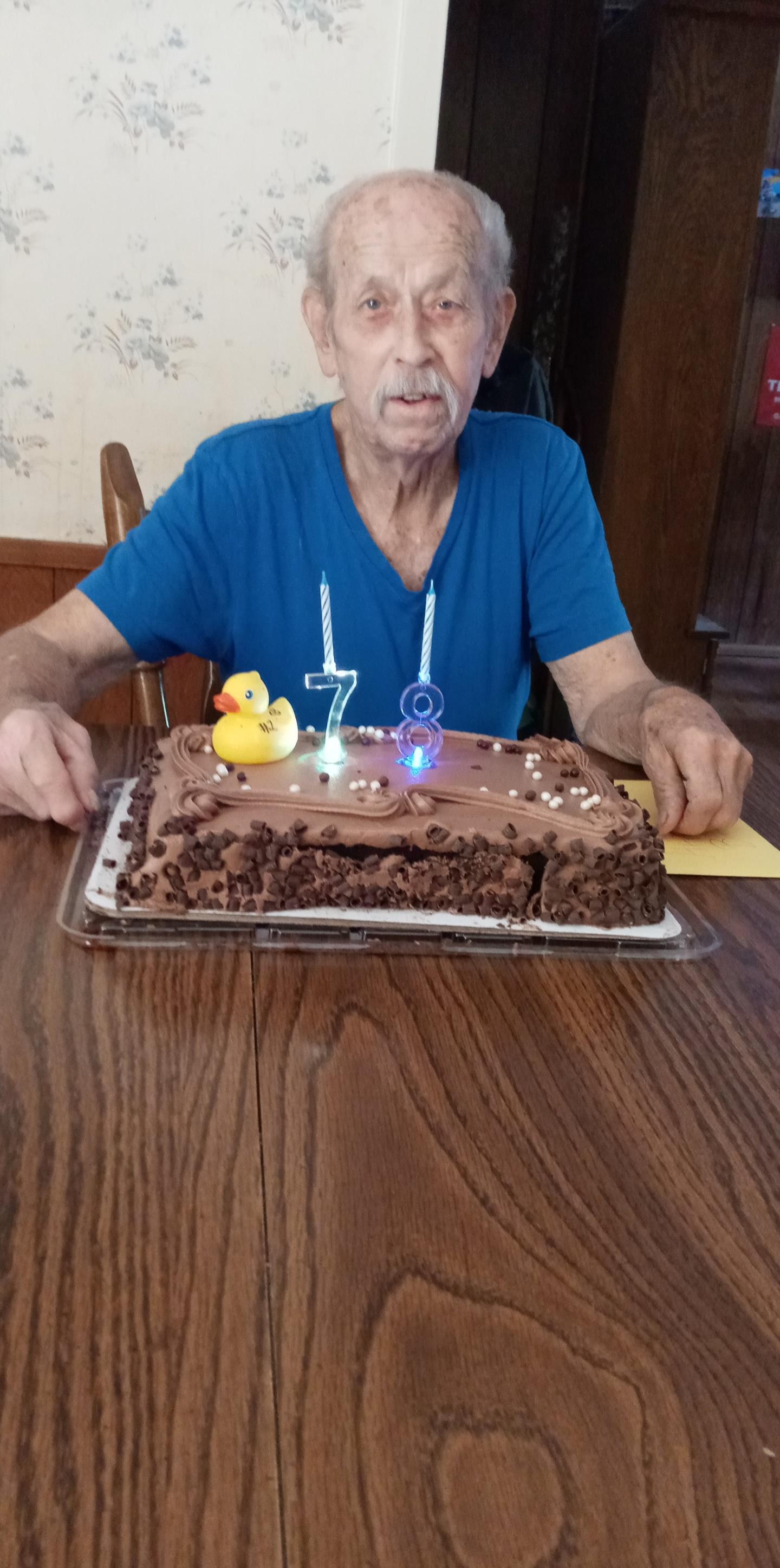 An elderly man sits at a table with a decorated chocolate cake, celebrating his birthday.