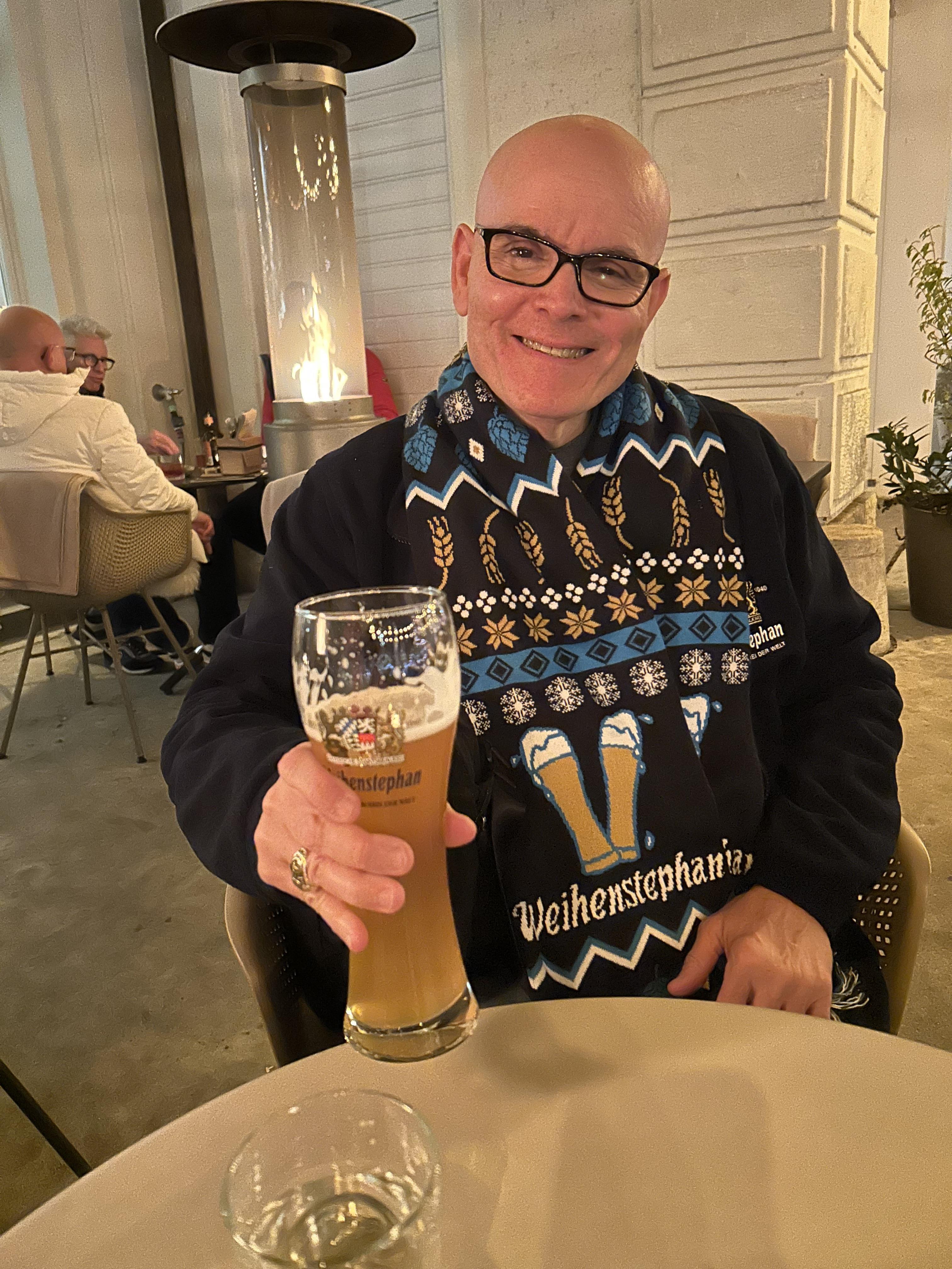 A man smiles and holds up a large beer in a stylish bar, showcasing his colorful sweater.