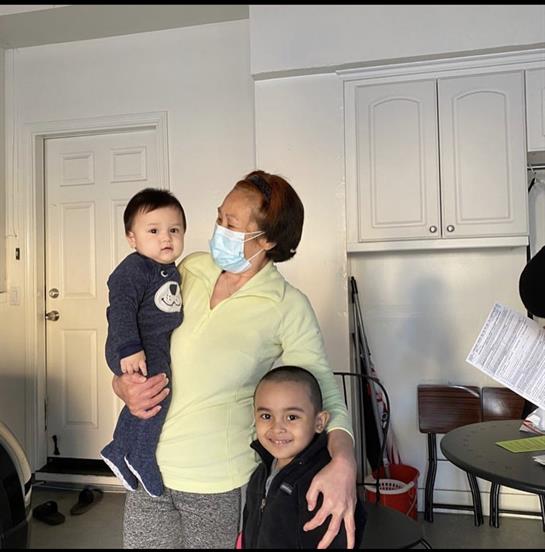 A woman in comfortable clothing holds a young child while another child stands close, smiling.