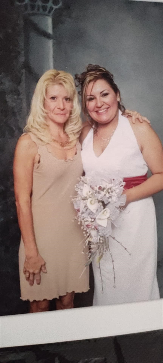 Two women pose together, one in a white dress holding a bouquet, in a decorated space.