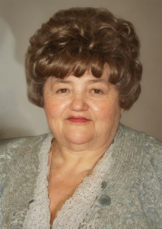 An elderly woman with curly hair smiles warmly while wearing a light cardigan indoors.