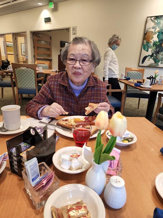 A senior woman smiles while eating breakfast with family in a cozy dining room.