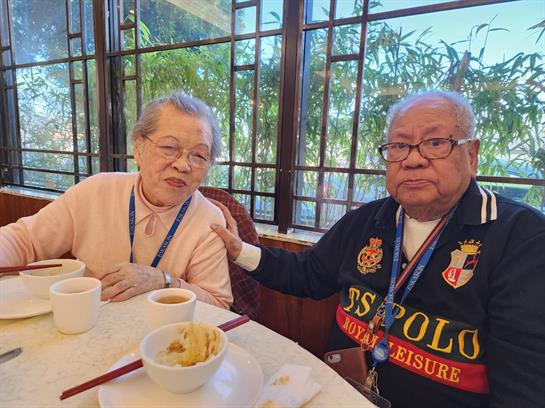 An elderly couple shares a meal and conversation in a welcoming restaurant surrounded by greenery.