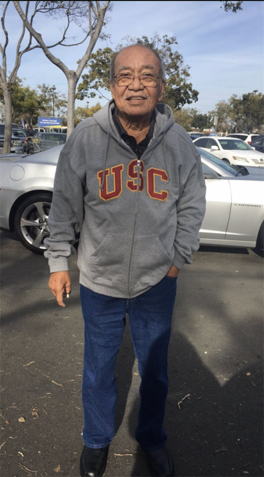 An elderly man wearing a USC hoodie stands in a parking lot on a sunny day.