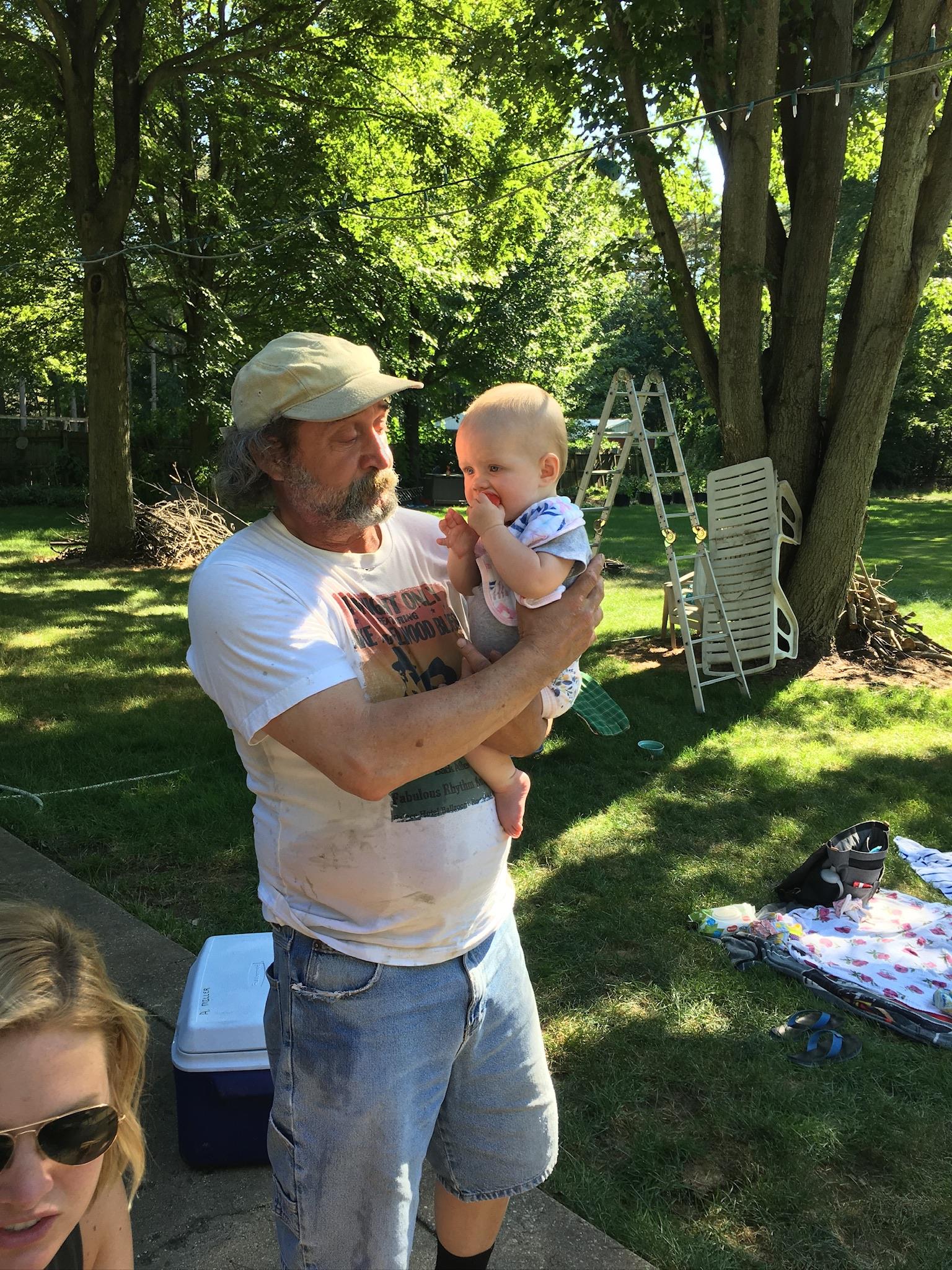 An adult with a hat carries a baby in a green space while enjoying the outdoors on a sunny day.