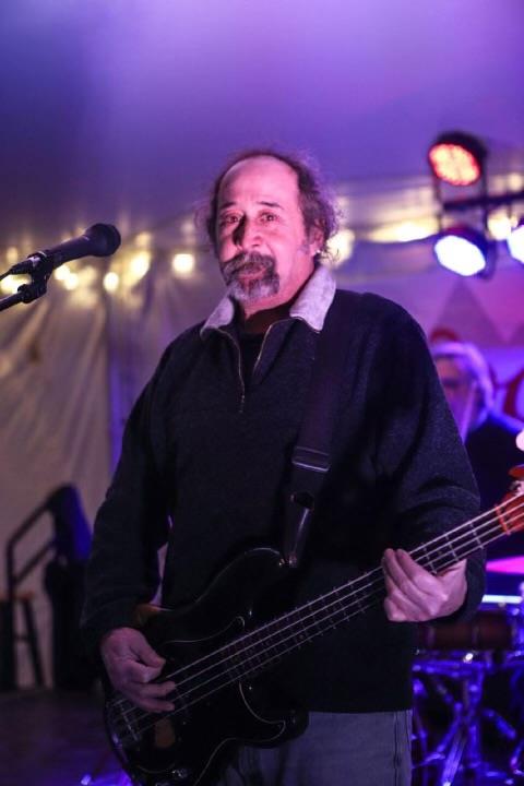 A musician plays the bass guitar while singing into a microphone at an outdoor event.