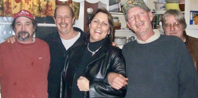 Five friends smile and pose together in a warm kitchen, enjoying each other's company late at night.