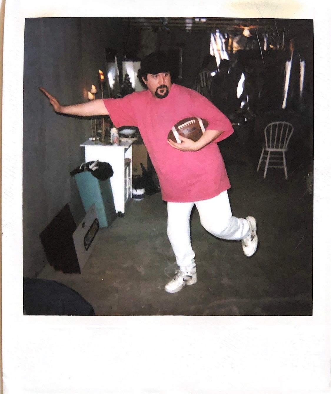 A man wearing a pink shirt and white pants poses joyfully while holding a football indoors.