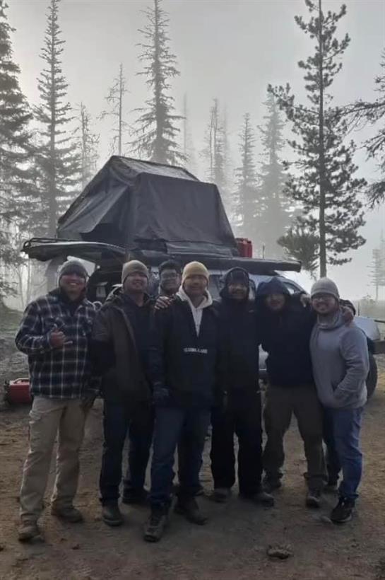 Friends gather around their campsite in the mountains, surrounded by tall pine trees and fog.