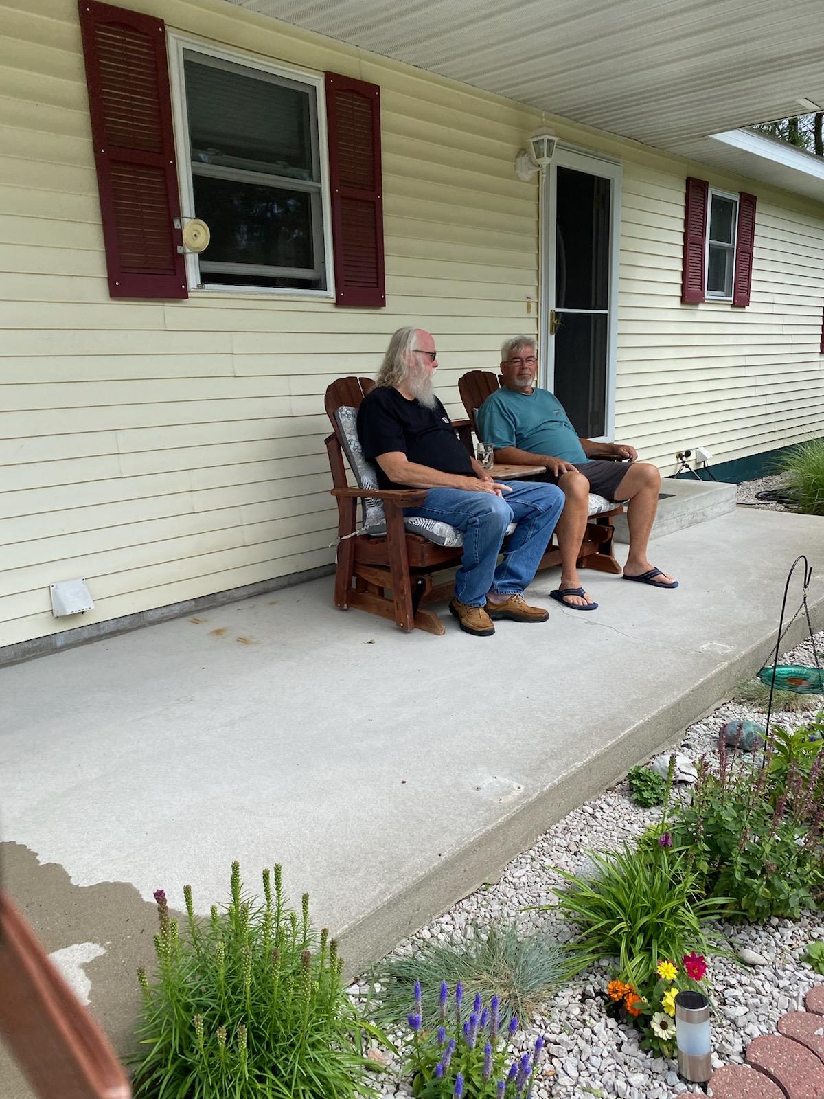 Two men sit comfortably on rocking chairs on a covered porch, enjoying the weather.
