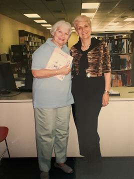 Two smiling women pose together at a library while holding certificates, celebrating a moment.