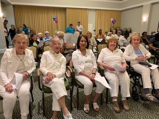 Several seniors sit together in white outfits, enjoying a lively community event in a hall.