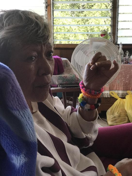An elderly woman sits in a cozy space, animatedly gesturing with bright bracelets on her wrists.