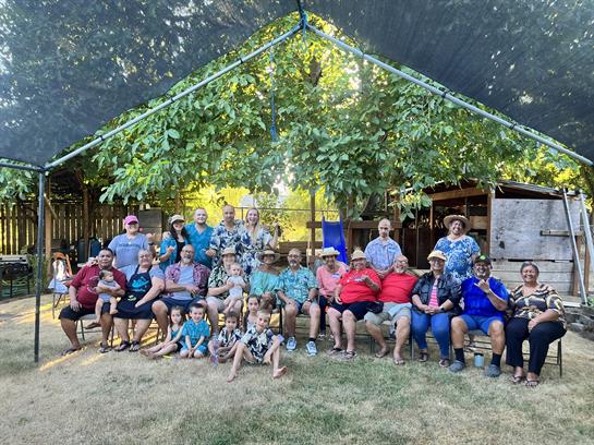 A large group of family members gathers happily under a shaded canopy, enjoying their time together.