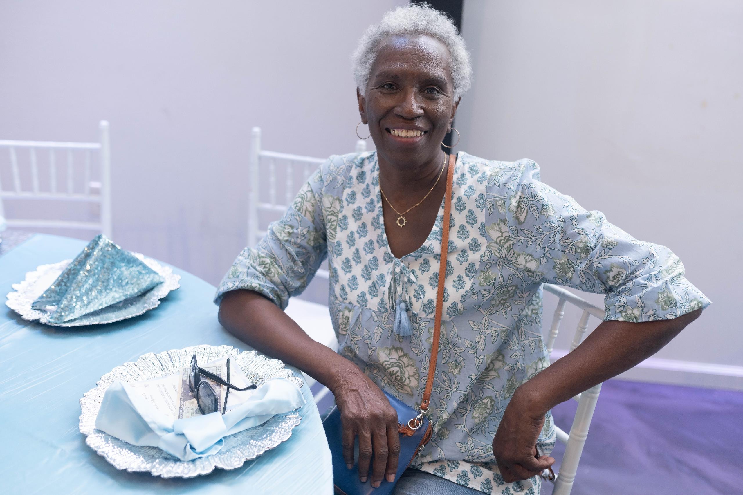 A cheerful elderly woman sits at a table adorned with blue decorations, ready for a celebration.