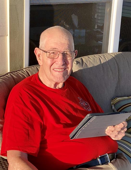 A man smiles while sitting comfortably on a porch with a tablet in hand during sunset.