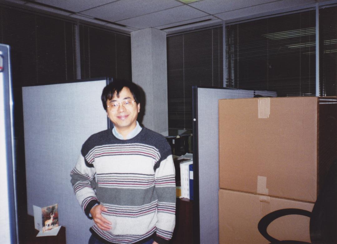 An individual stands in a dimly lit office space, smiling while surrounded by cubicles and boxes.