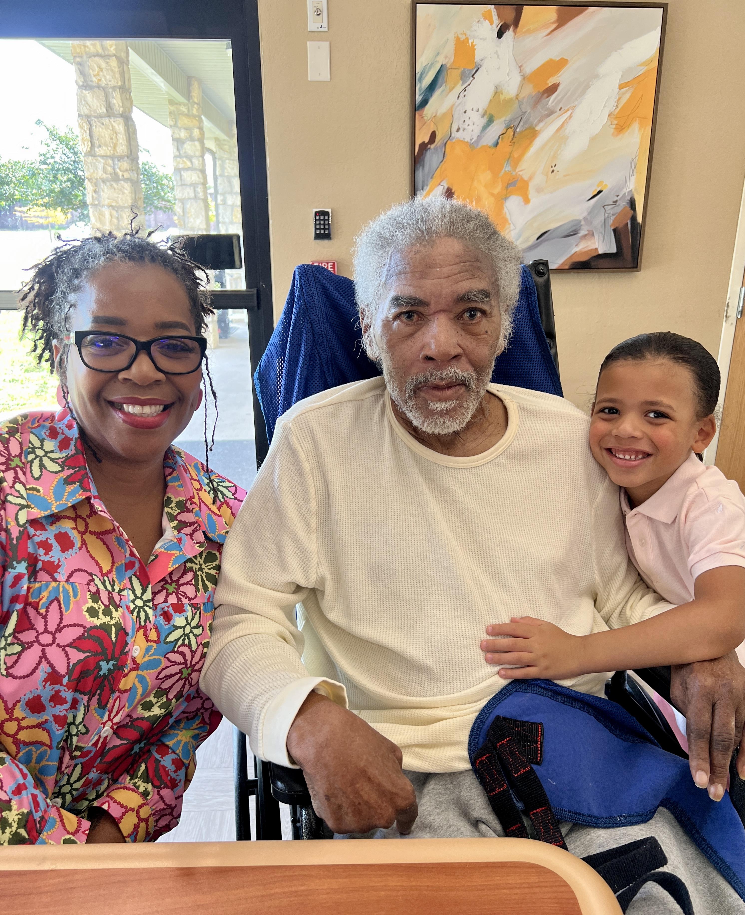 A grandmother and grandson joyfully pose with their grandfather in a nursing home setting.