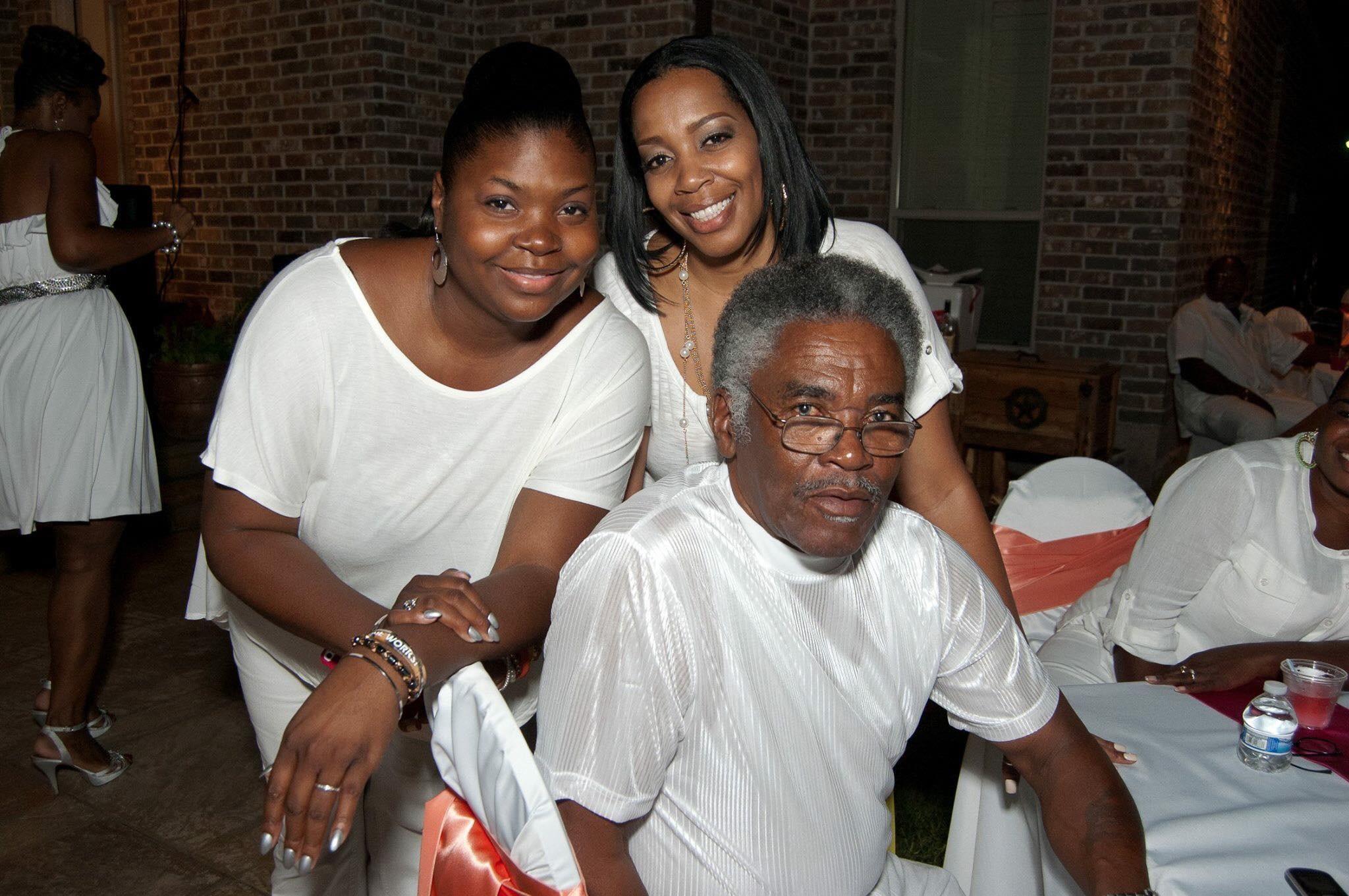 Two women joyfully pose with an older man during a family gathering in evening attire.