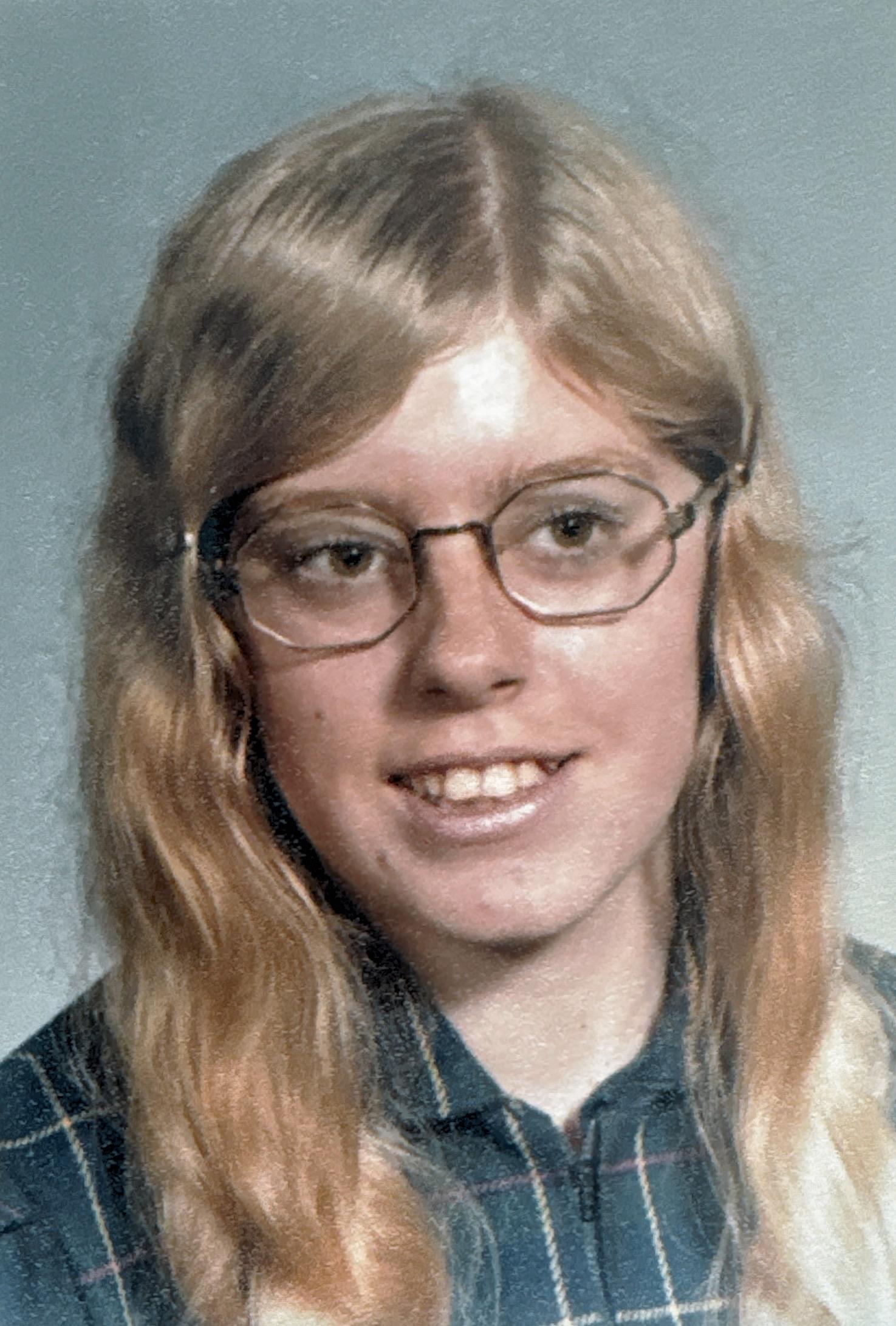 A young girl with long blonde hair smiles warmly, wearing glasses and a plaid shirt.