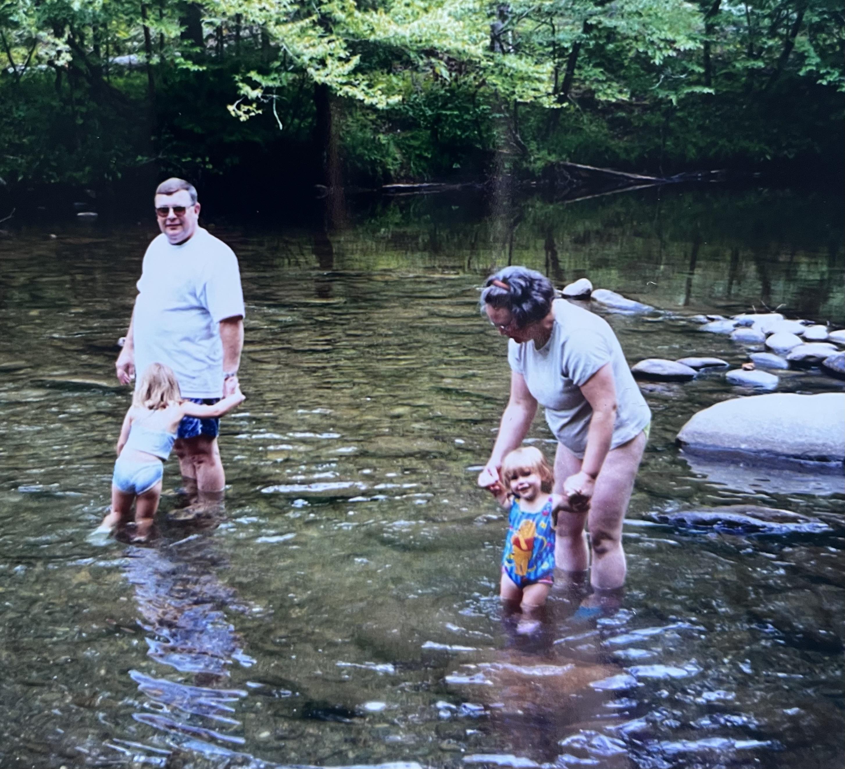 Adults and children splash joyfully in a shallow river under warm sunlight surrounded by trees.