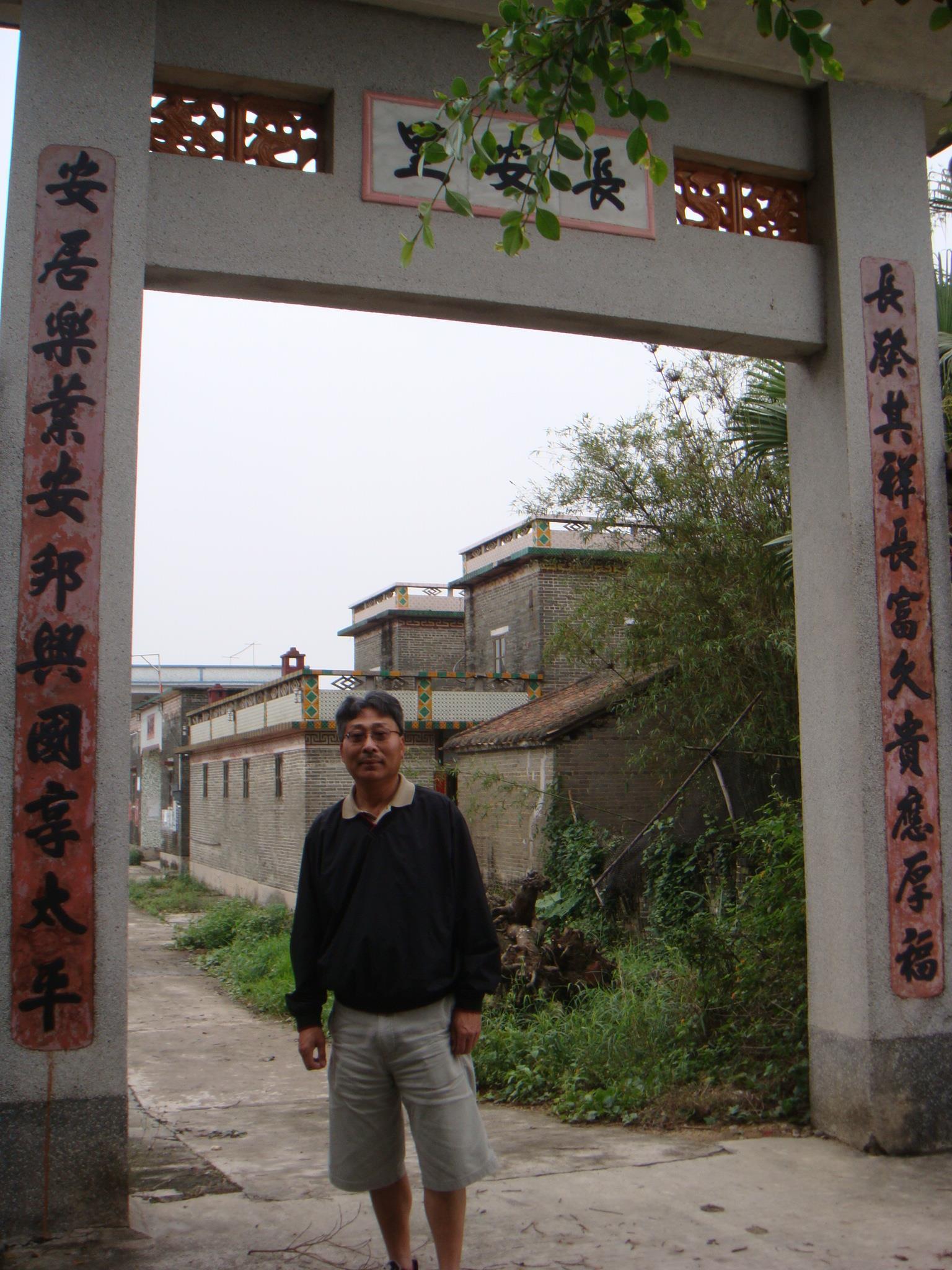 A man poses at the entrance of a rural village, surrounded by an archway and greenery.