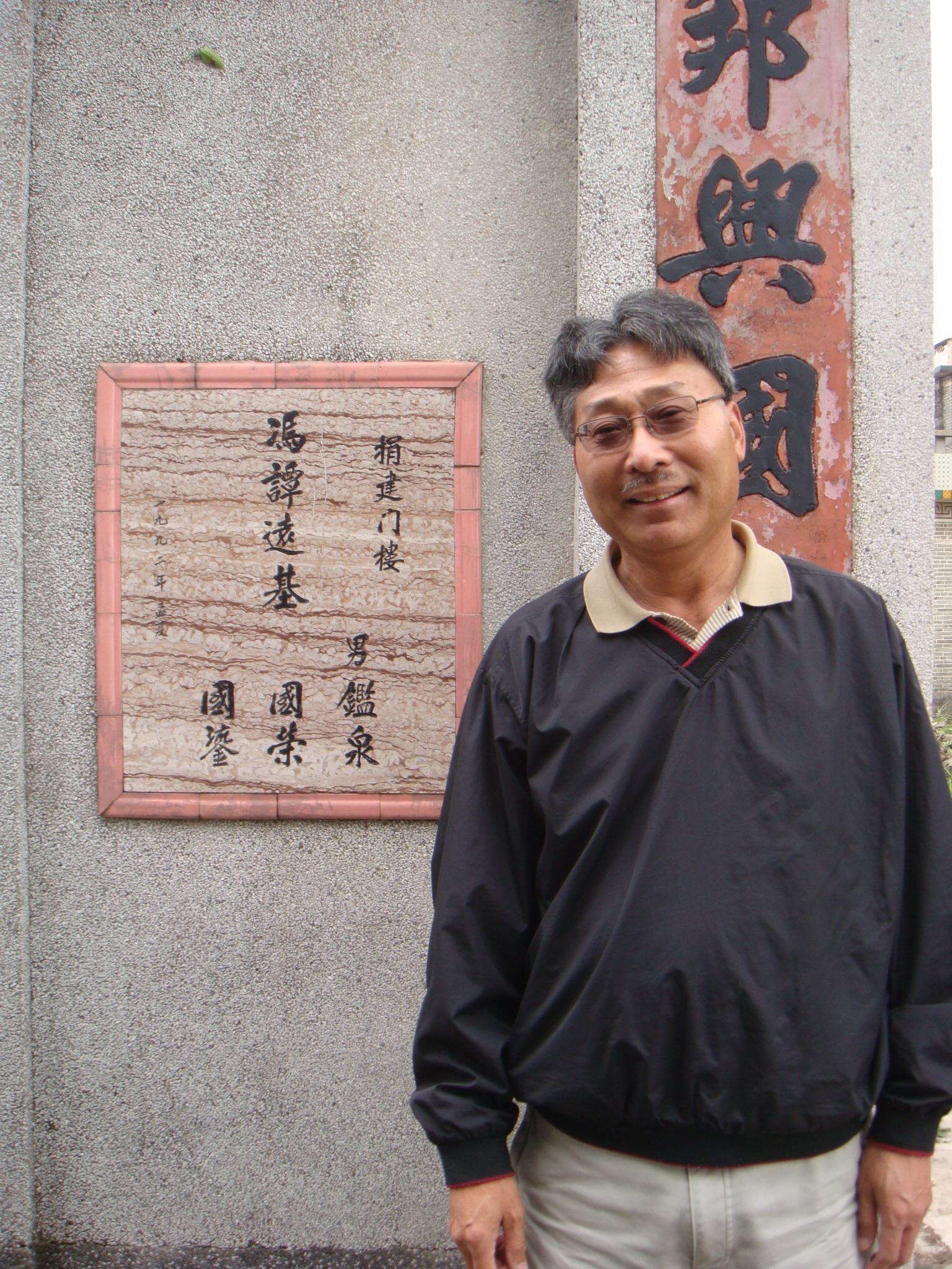 A man stands smiling near a stone structure with Chinese inscriptions in an urban setting.