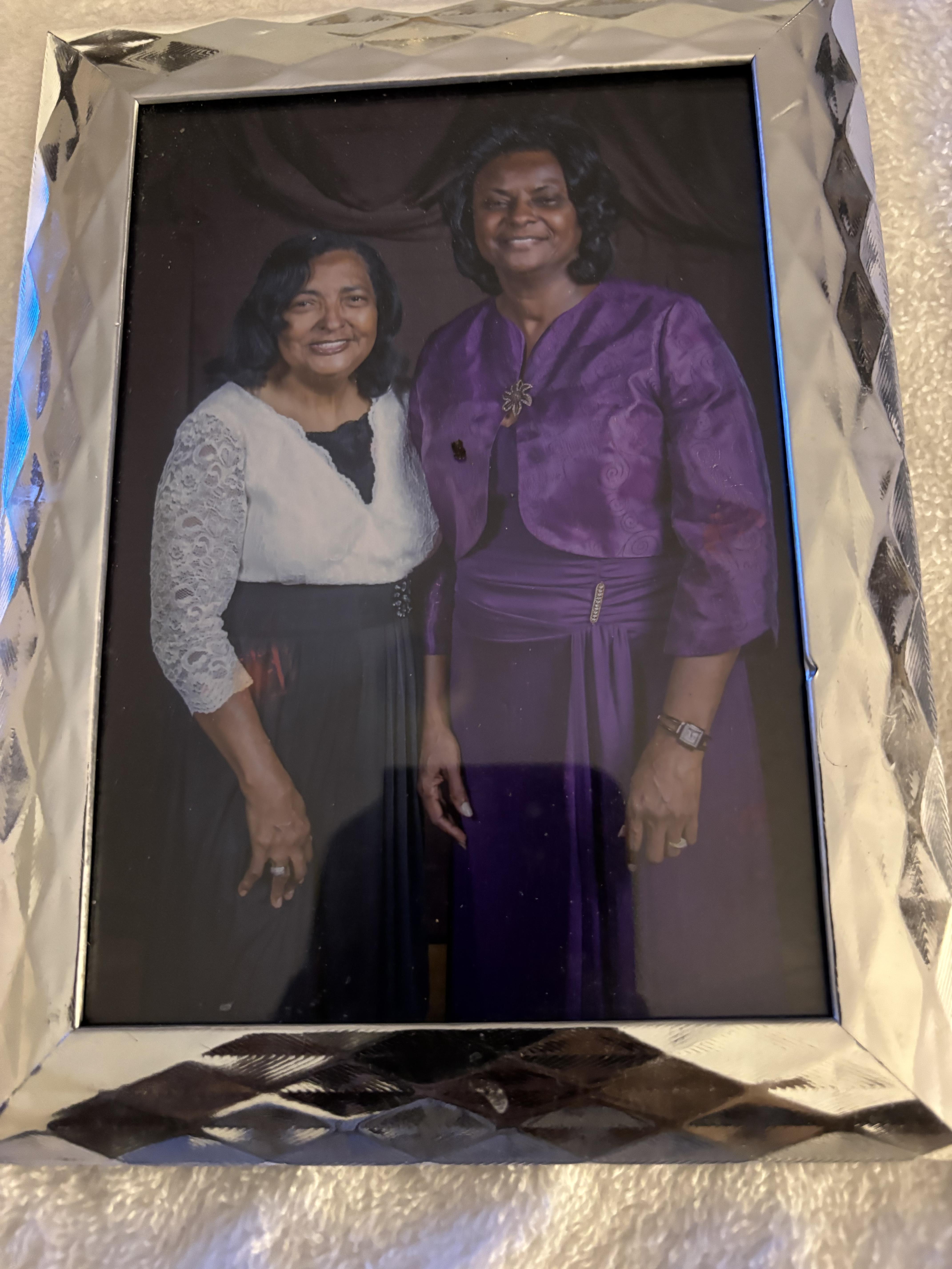Two women stand side by side, smiling warmly while dressed elegantly in complementary outfits.