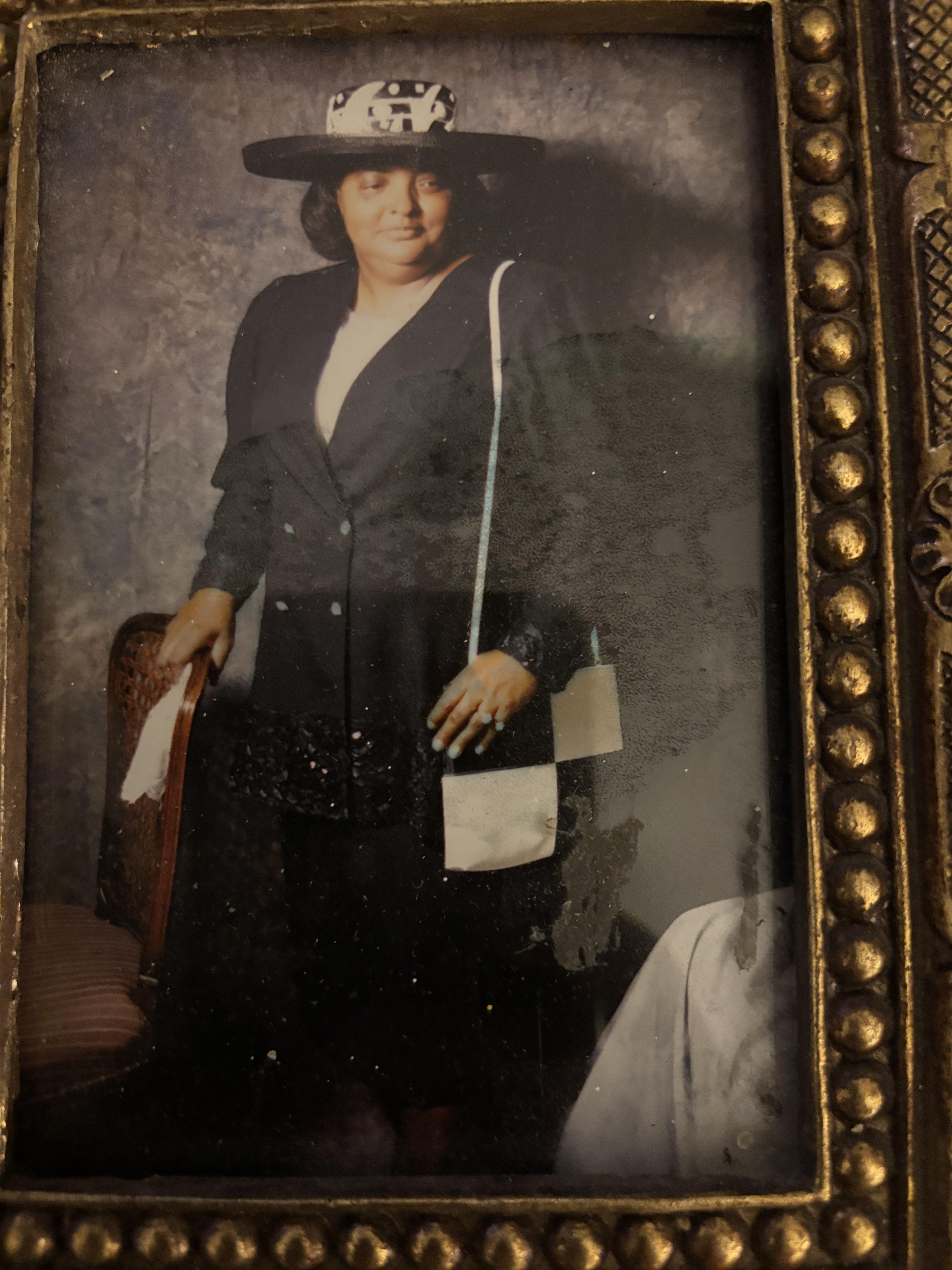 A woman wears a stylish hat and dark outfit, standing confidently in a vintage room.