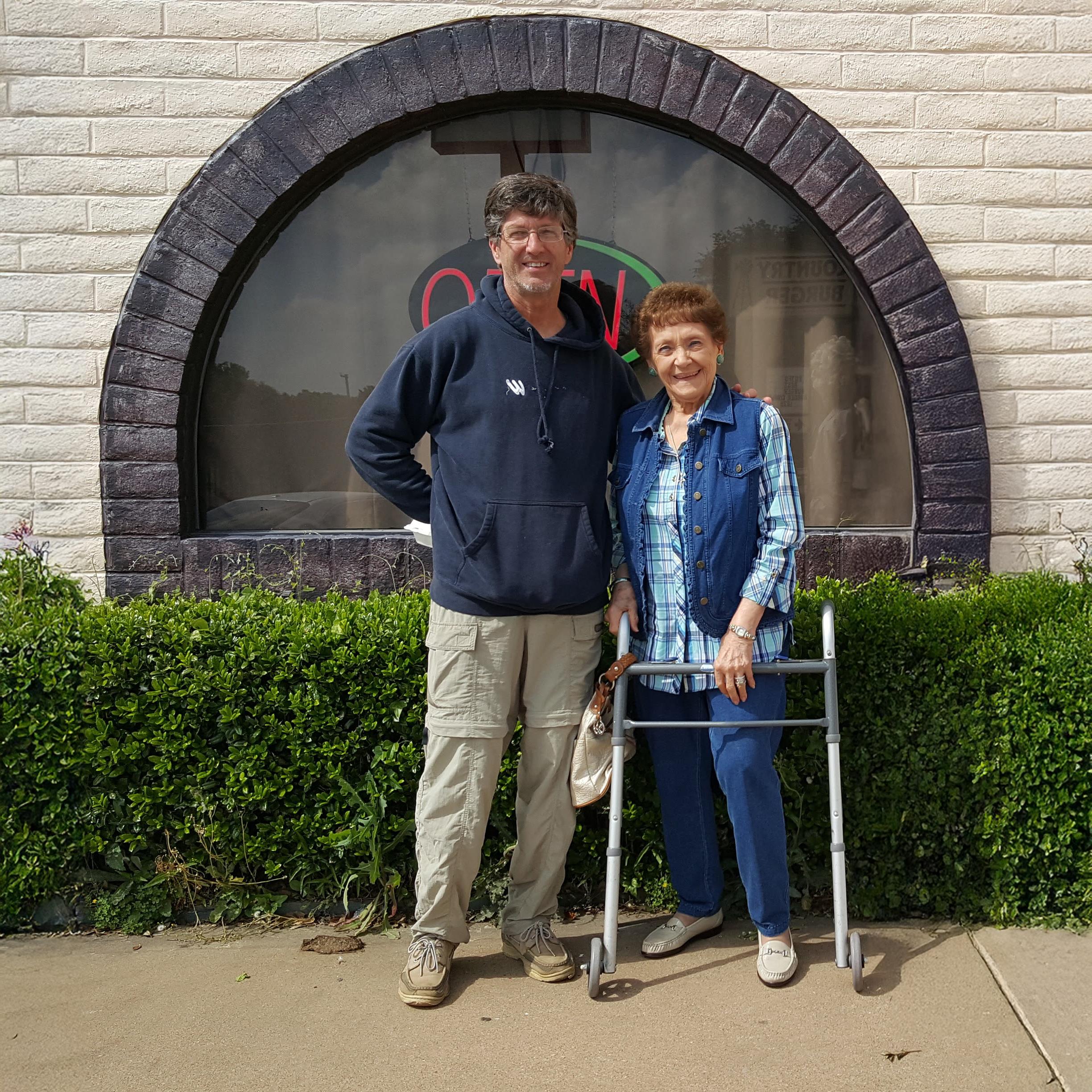 Two people smile as they stand outside a building, surrounded by plants and bright sunlight.