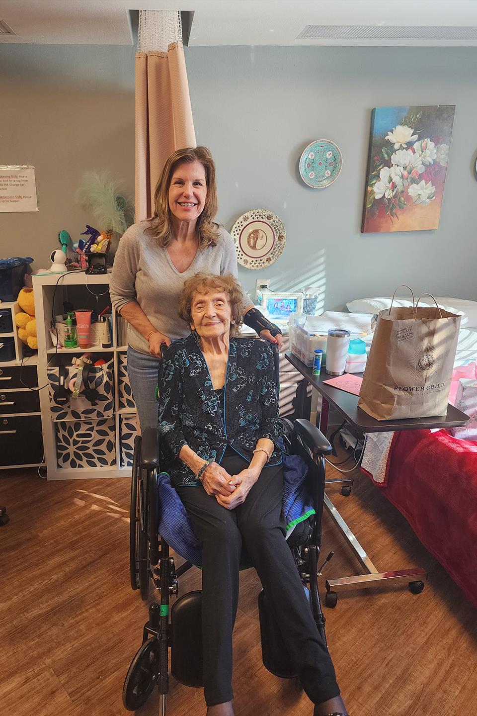 A caregiver stands behind an elderly woman seated in a wheelchair, both smiling happily.