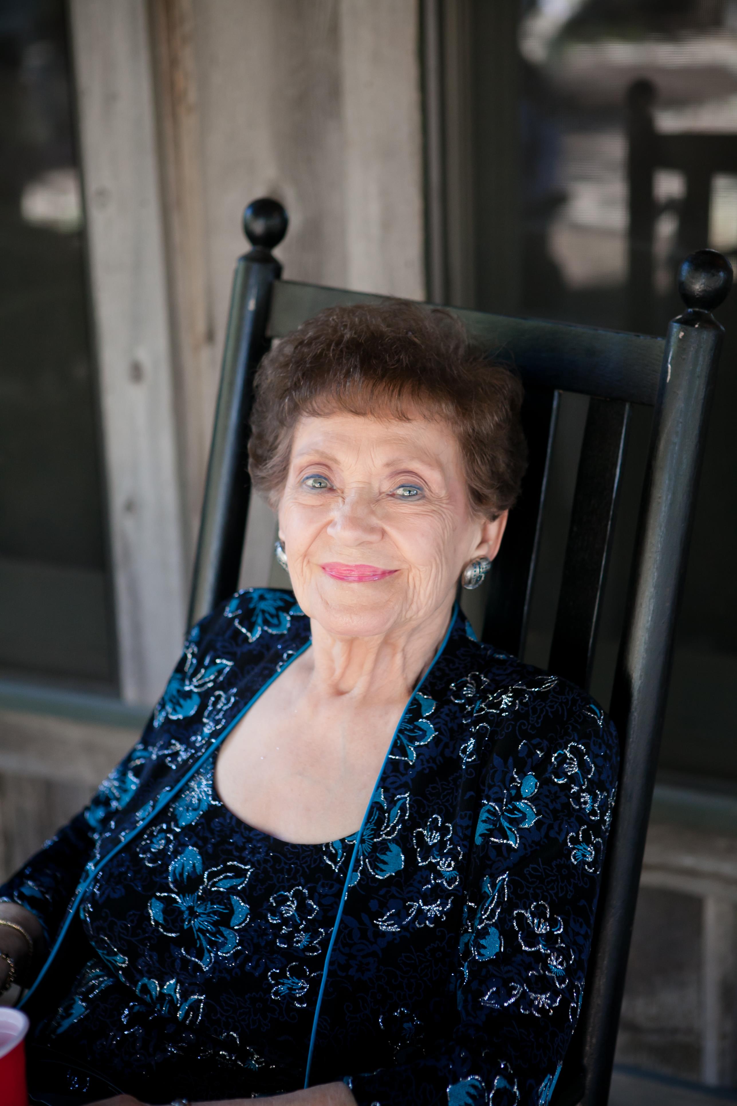 An elderly woman relaxes in a rocking chair, smiling in the warm sunlight on the porch.