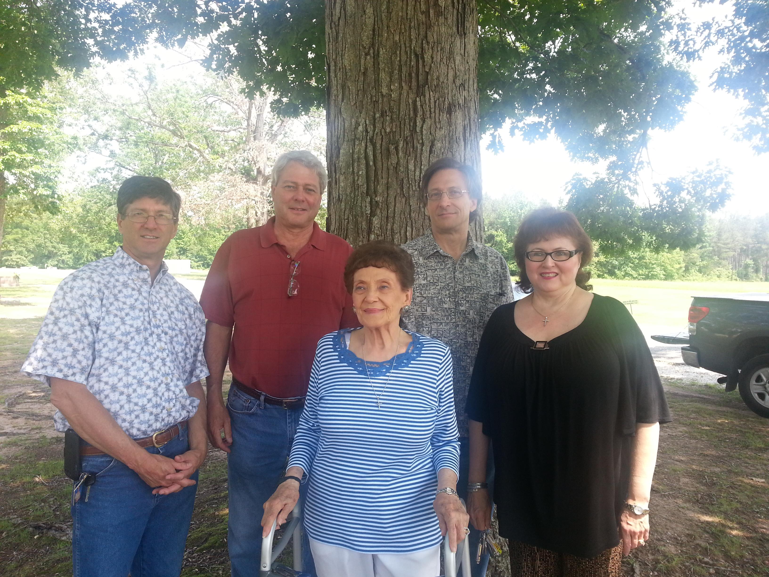 Five family members stand together near a large tree in a park on a bright day.