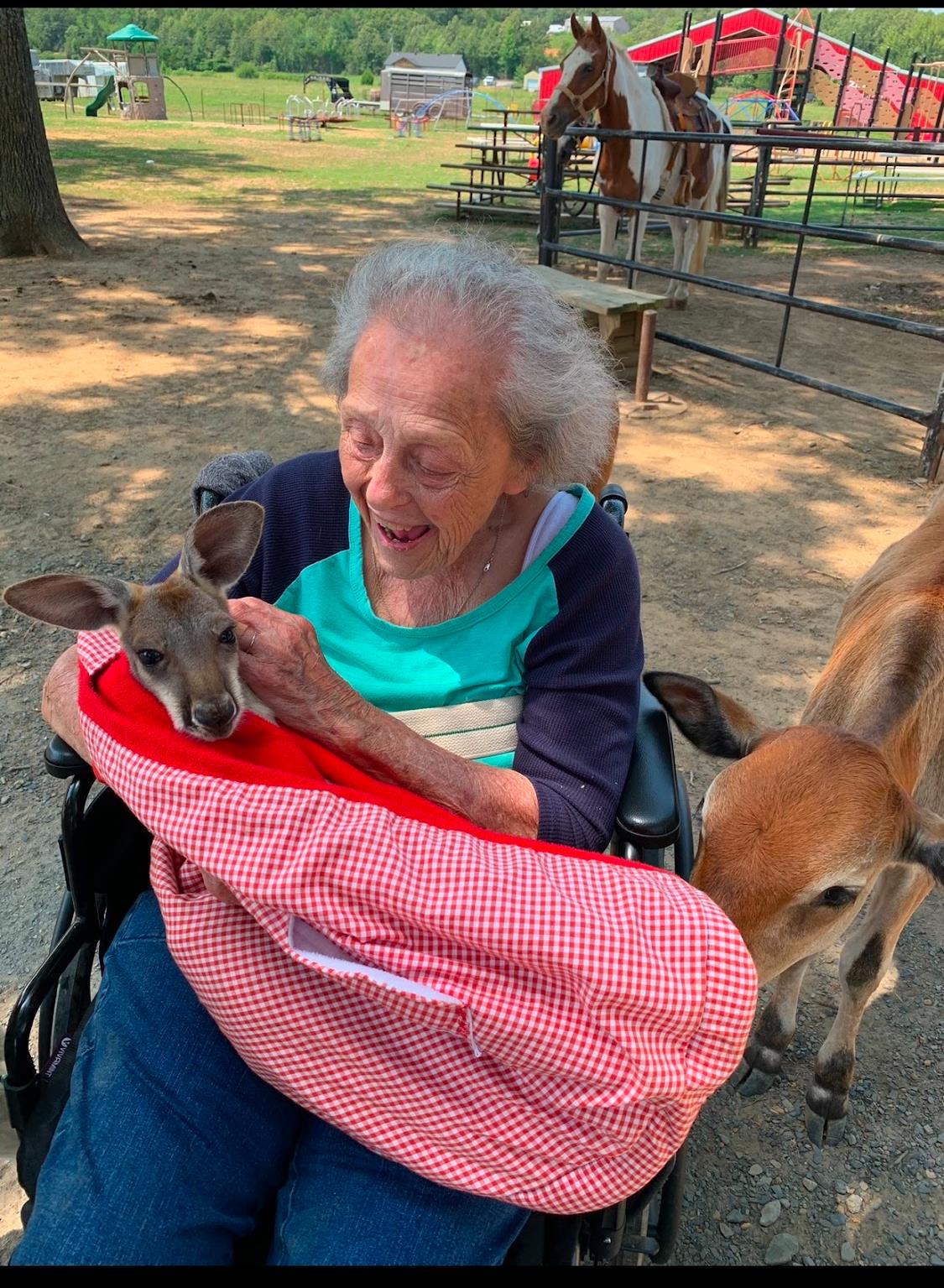 A senior woman in a wheelchair smiles as she gently caresses a baby deer in her lap.