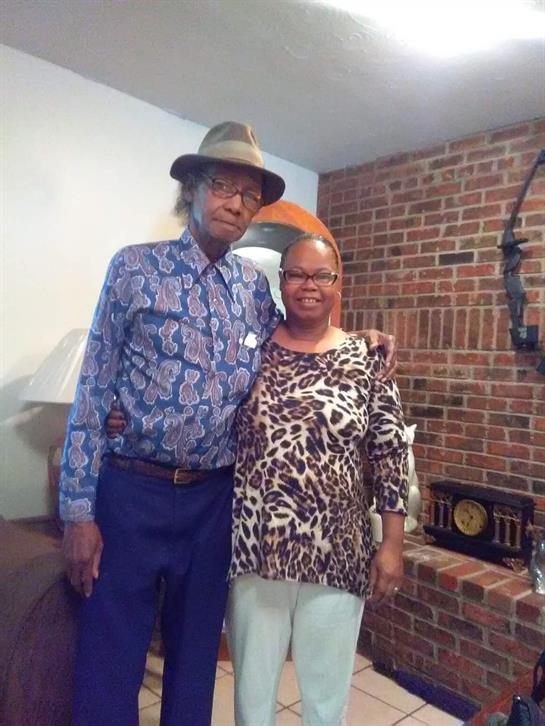 Two smiling individuals stand together in a warm living space, sharing a joyful moment.