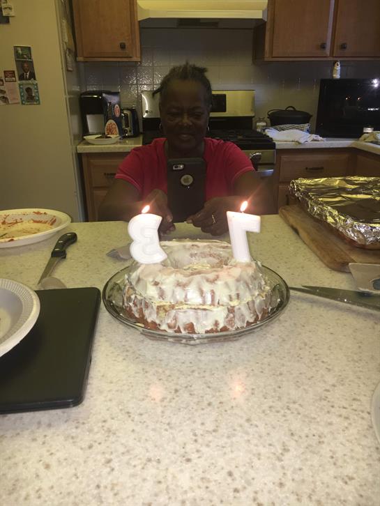 A person celebrates their birthday with a candle-topped cake in a cozy kitchen.