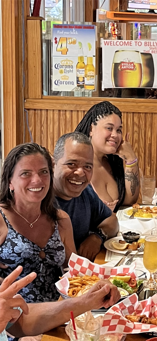 A group of three friends smiles while seated at a busy restaurant table, sharing food and drinks.