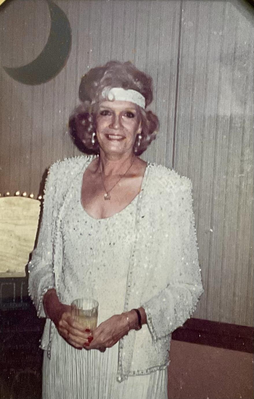 An elderly woman dressed in elegant attire stands smiling while holding a drink at a gathering.
